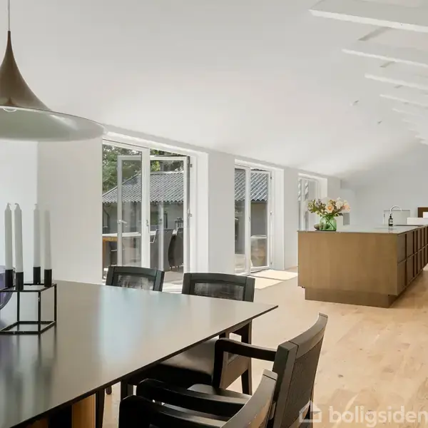 Spisebord med fire stole står i lys stue med store vinduer. Et moderne køkken med en ø er i baggrunden. Rummet har lyst trægulv og hvidt loft med synlige bjælker.