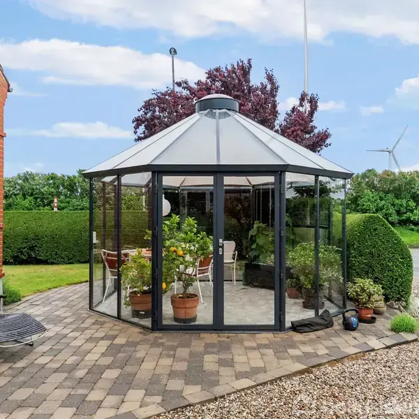 Et glashus står på en brostensbelagt terrasse, omringet af grønne planter og havemøbler, ved siden af en murstensbygning med hække og vejrtræ.