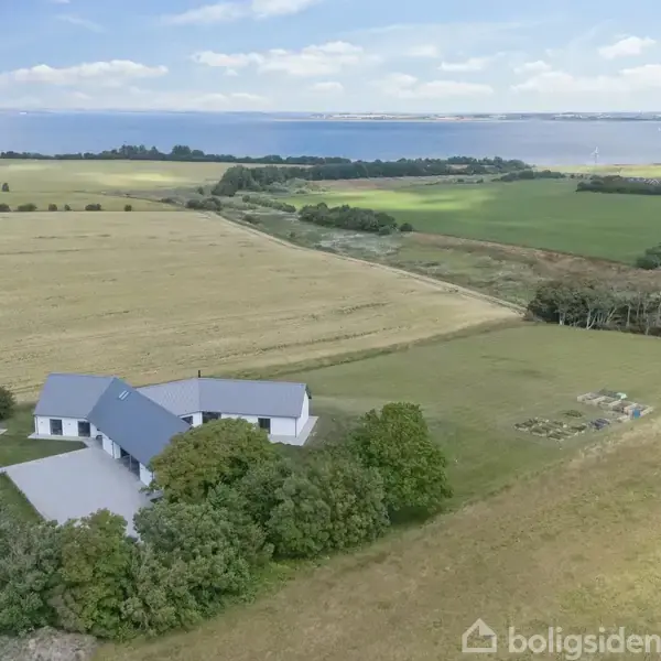 En hvid bygning med gråt tag ligger blandt træer midt i åbne marker. I baggrunden strækker en blå fjord sig mod horisonten under en let skyet himmel.