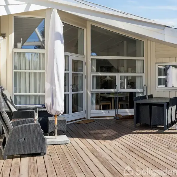 En terrasse med trædæk er udstyret med sorte havemøbler og en stor parasol, placeret foran moderne, store vinduer til et hus. Haven ligger i et solrigt udemiljø.