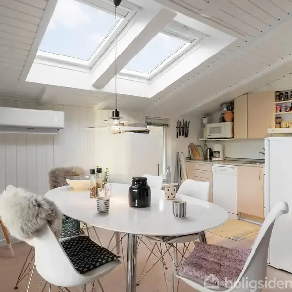 En lys spisestue med et rundt bord omkranset af stole, placeret under ovenlysvinduer. Køkkenredskaber, krydderier og køkkenapparater fylder hylderne i det bageste køkkenområde. En lille dansk flag står i hjørnet.