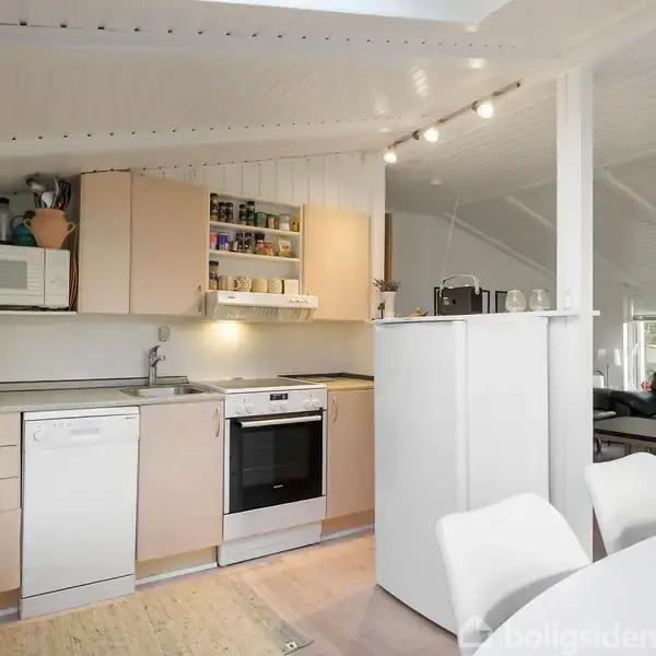 Et moderne køkken med opvaskemaskine og ovn under beige skabe; en mikrobølgeovn står til venstre. I baggrunden ses en stue med sofa og sofabord i et lyst rum.