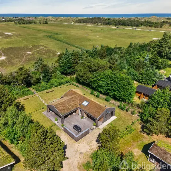 Et moderne etplanshus med græsbelagt tag vises omgivet af frodig skov og åbne marker. En stor terrasse med bord og stole er foran huset, som er beliggende i naturskønne omgivelser.