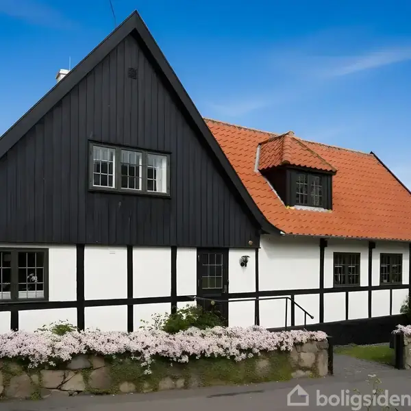 Hus med sort træværk og hvid facade, rødt tegltag; beliggende ved kysten omgivet af blomstrende buske og stengærde. Himmel og hav ses i baggrunden.