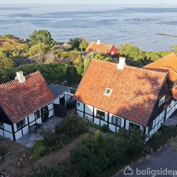 Traditionelt bindingsværkshus med rødt tegltag ligger ved kysten omgivet af grønt landskab og havudsigt. Flere lignende bygninger er synlige i baggrunden nær vandkanten.