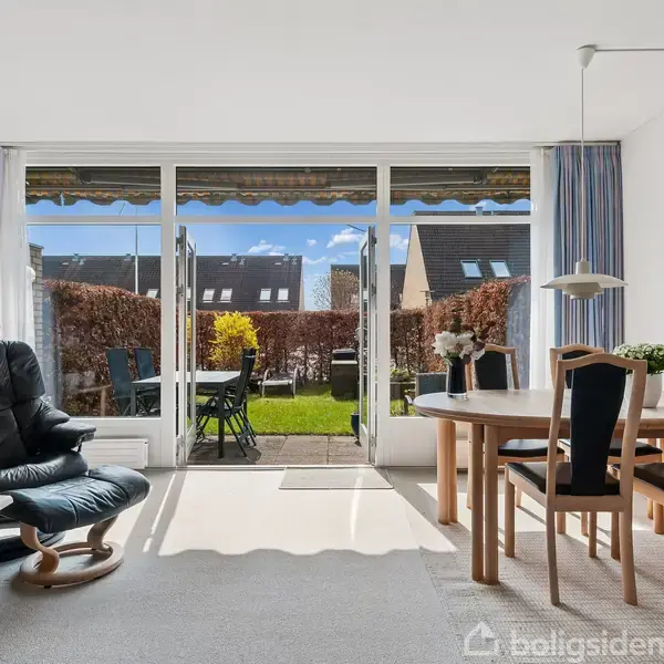 Stue med lænestol og spisebord; store vinduer åbner ud til en have med terrasse og hække. Blåt og hvidt interiør, lys træmøbler og en maleri hængt på væggen.