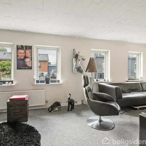 Stue med sorte lædersofaer og en drejestol ved siden af et firkantet sofabord, dekoreret med kunstplakater og skulpturer på væggene, lysindfald fra fire vinduer.