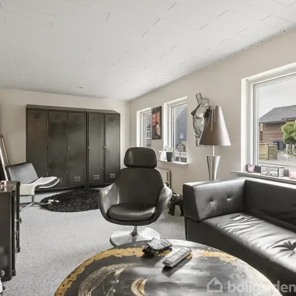 En stue med en sort lædersofa og en lænestol ved et sofabord. Store vinduer lyser rummet op, mens et skab står bagtil. En lampe og dekorationer er også synlige.