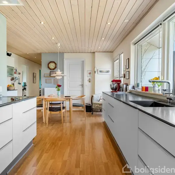 Køkkenet har hvide skabe og grå bordplader, udstyret med vask og køkkenudstyr. I baggrunden står et spisebord med fire stole. Lyse trægulve og lyse vægge skaber en lys atmosfære.