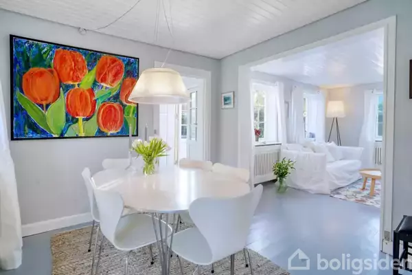 En rund, hvid spisestuebord med seks stole står i et lyst rum. Over bordet hænger en lampe, og en farverig maleri med røde blomster pryder væggen.