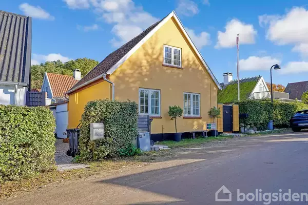 Gul murstenshus står stille på en solrig gade, omgivet af hække og flere huse i baggrunden. To vinduer vender mod gaden, og der er en flagstang ved siden af.