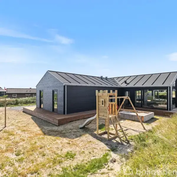 Et moderne, sort træhus står stille i et åbent landskab med sand og græs. Ved siden af huset er der en lille legeplads med gyngestativ. Øverst ses teksten: "Modelfoto".