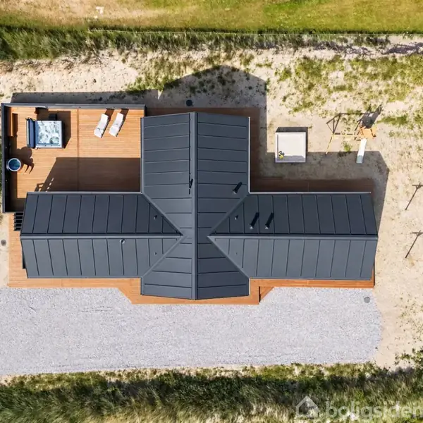 T-formet sort taghus set ovenfra, omgivet af sand og buske. En træterrasse med møbler er synlig på venstre side. Tekst: "Modelfoto" i en cirkel.