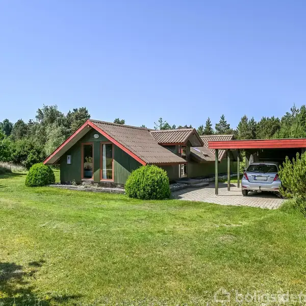 Hus med rødt sadeltag står i en grøn have. En bil holder parkeret under en carport ved siden af huset. Omgivende skov ses i baggrunden under blå himmel.