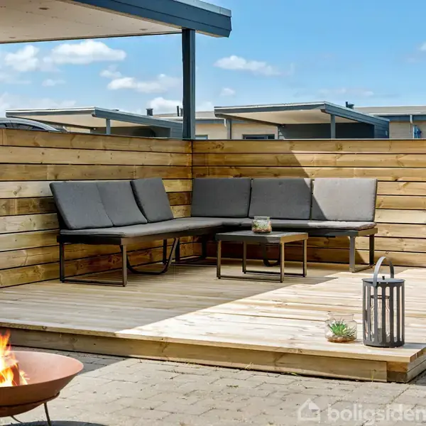 Terrasse med gråt sofasæt placeret i hjørnet på trædæk, inden for rammerne af et hegn. En lille bordlampe og plante på bordet. I forgrunden ses en ildskål på stenbelægning.