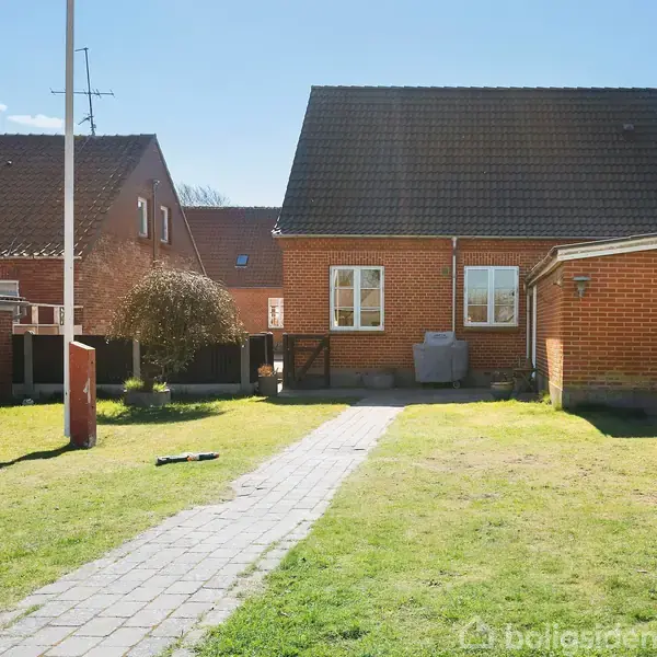 Rødt murstenshus med mørkt tag står bag en græsbeklædt have. Et flisebelagt stisystem fører fra forgrunden mod huset. Haven er omkranset af buske og træer.