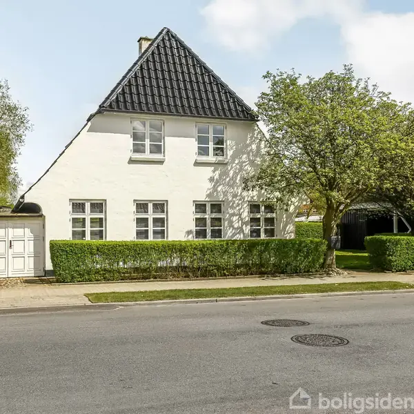Hvidt hus med sort tag står stille omgivet af grønne hække og træer. Det er placeret ved en vej i et beboelsesområde.