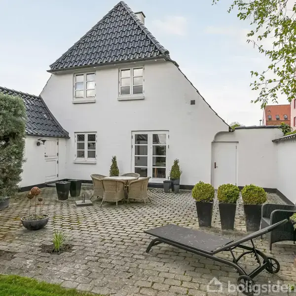 Hvide hus med sort tag, omgivet af en stenbelagt terrasse. Havemøbler står i midten, krukker med planter rundt om. En liggestol til højre, grønne buske til venstre.