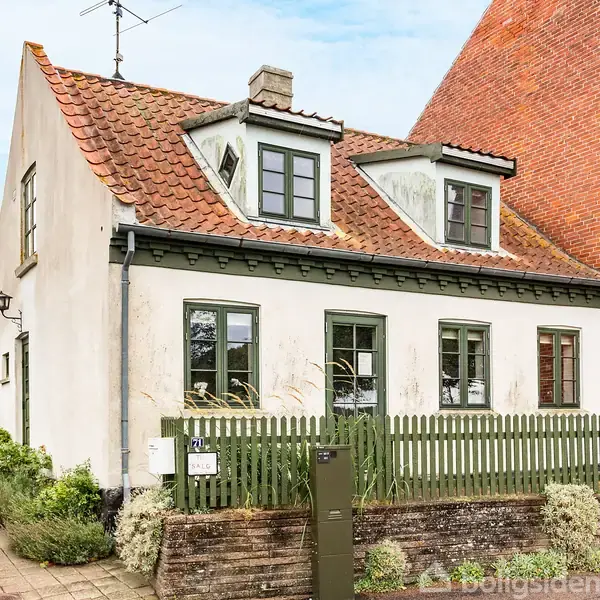 Et hvidt hus med rødt tegltag står bag et grønt stakit. Huset har to kviste, tre vinduer, og er omgivet af planter i en bymæssig setting.