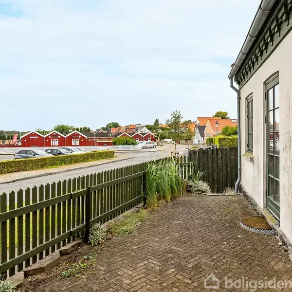 Et hus med hvide vægge og grønne vinduer står ved en brostensbelagt sti, indhegnet af et grønt plankeværk. I baggrunden ses røde bygninger og veje.