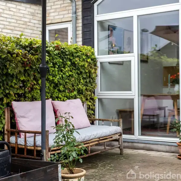 Bænk med lyserøde puder står på en terrasse nær et vindue. En grøn hæk omgiver området, og en vandkande og planter pynter omgivelserne.