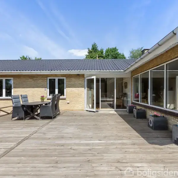 Hus med lysegule mursten og store vinduespartier åbner sig mod en stor træterrasse. Terrassen har et spisebord med stole og blomsterkrukker. Grønt træområde i baggrunden.
