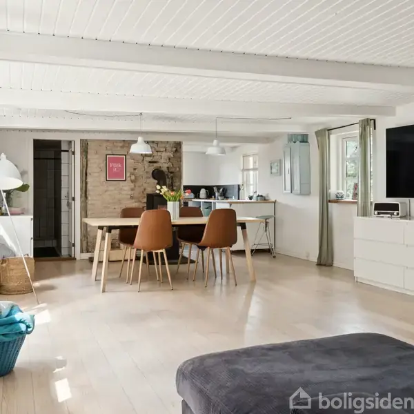 Stue med spisebord og stole i centrum, pejs og vindue i baggrunden. Moderne indretning med lys trægulv og hvidmalede lofter. Adjacent køkken og stor, monteret fladskærm.