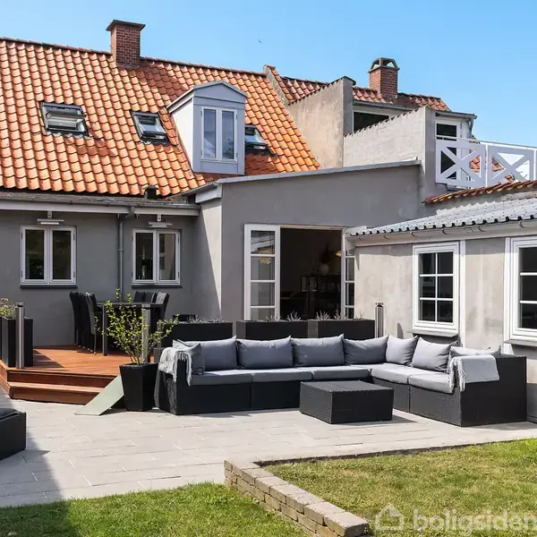 En grå murstenshus med rødt tegltag har en terrasse med en U-formet sofa. Terrassen har planter og et græsareal i forgrunden.