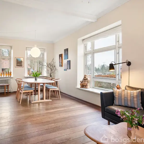Lænestue med sort sofa, træbord og blomster på en lys trægulv. Rundt spisebord med stole ved store vinduer. Hyggelig atmosfære med malerier og planter i hjørnet.