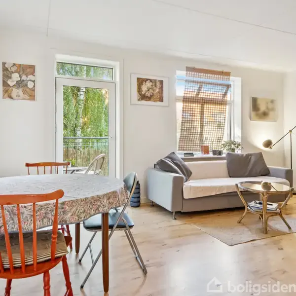 En stue med en rund spisebord med stole, en hvid sofa med puder, og en guitar i et stativ. Rummet er lyst med naturligt lys fra vinduer.