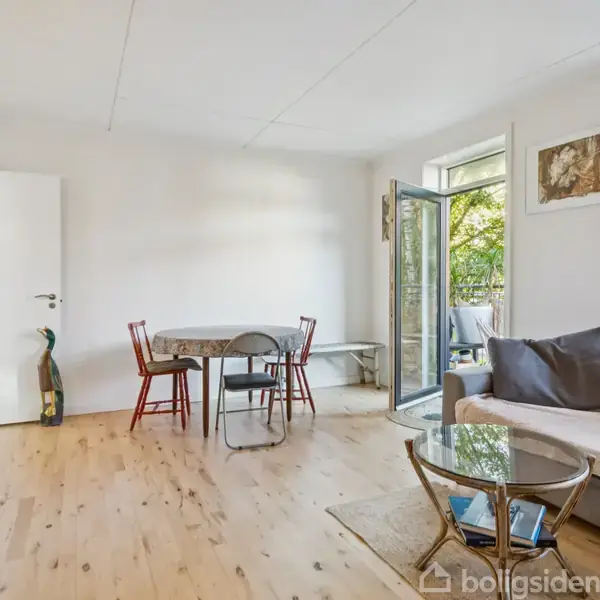 Stue med sofa og glasbord, åben terrassedør, træbord med stole, guitar mod væg, og maleri. Rummet er lyst med trægulv, hængende lampe og udsigt til grønne træer udenfor.