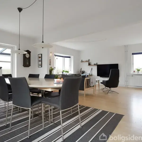 Spisebord med sorte stole står på en stribet tæppe i en lys stue. Rummet har store vinduer, en vægmonteret TV og moderne indretningselementer.