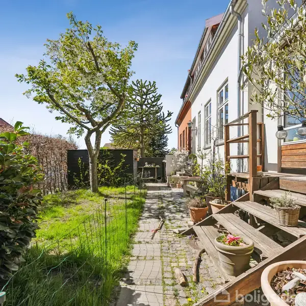 Trappe fører op til terrasse foran hus omkranset af blomsterkrukker. En grøn have med et træ og hækkede buske ses i baggrunden under blå himmel.