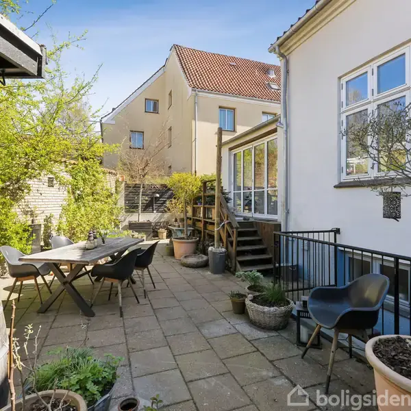 Terrasse med fliser har havemøbler og krukker med planter. Et hus med store vinduer er til højre, og trappen fører op til et hævet dæk. Træer omkranser området.