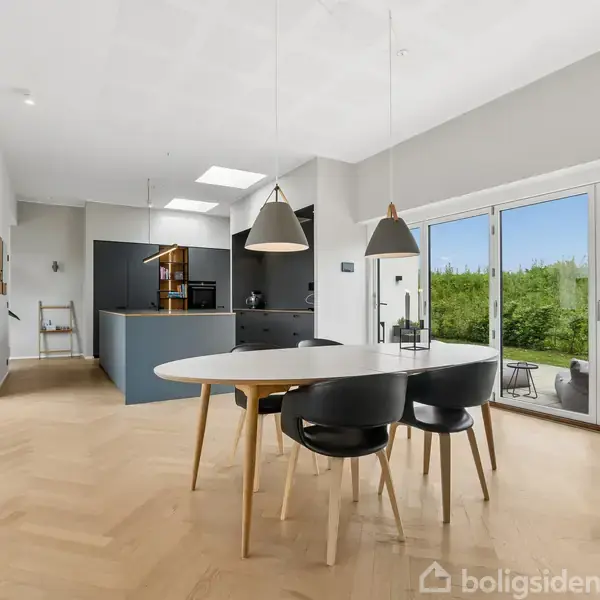 Spisebord med sorte stole står i et lyst køkken-alrum. Store vinduer til højre viser en terrasse med udsigt til en grøn hæk. Køkkenet har hvide og mørke skabe.
