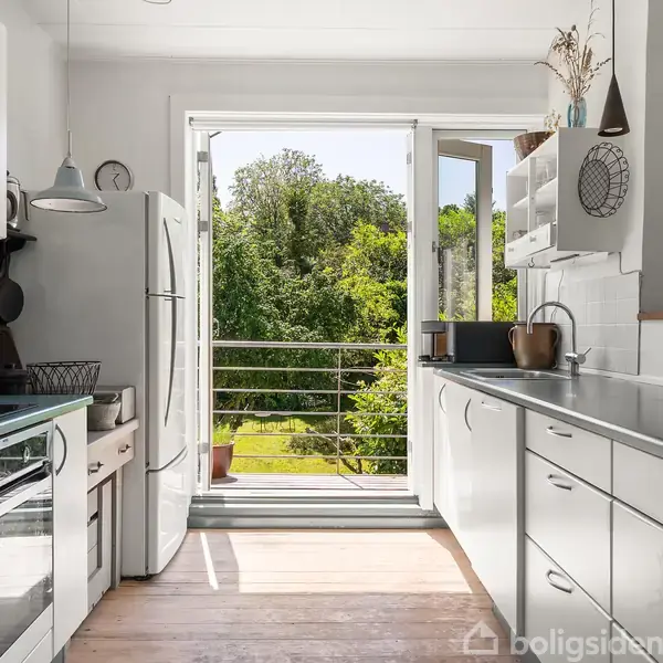 Et hvidt køkken åbner ud mod en grøn terrasse gennem franske døre. Lyse træbordplader og stålgreb dominerer, mens naturens farver trænger ind gennem de åbne døre i baggrunden.