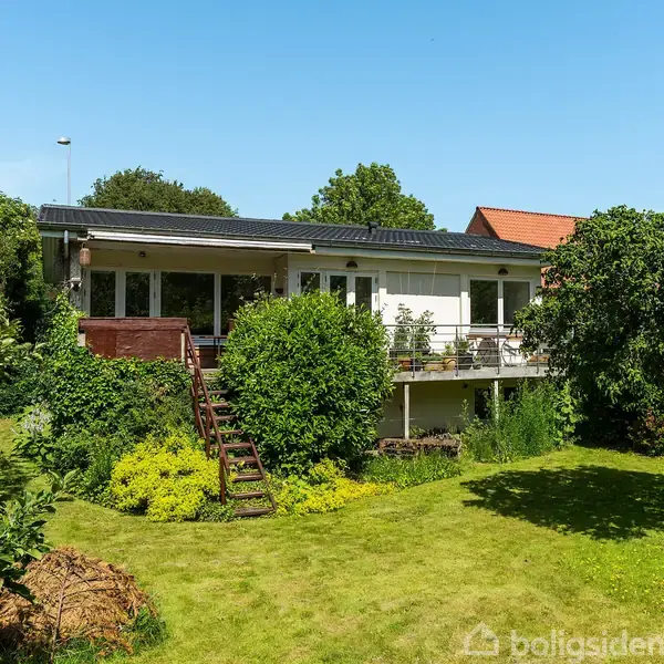 Huset fremstår som en hvid etplans villa med terrasse, omkranset af frodige buske og træer. En velplejet græsplæne strækker sig foran villaen i en solrig have.