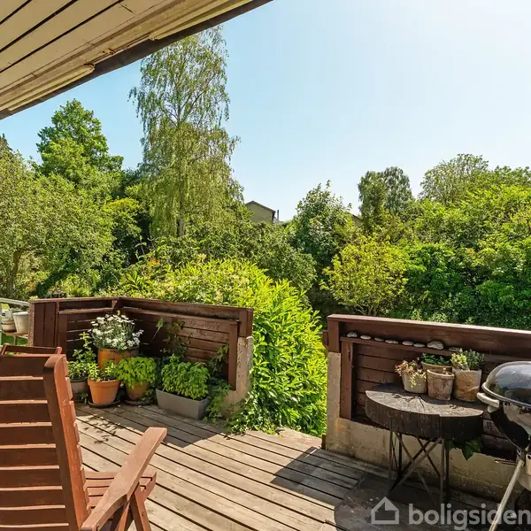 En træterrasse med havemøbler og en grill står i solen. Omkring er der tæt grøn beplantning og træer, der giver en fredfyldt, naturskøn atmosfære.