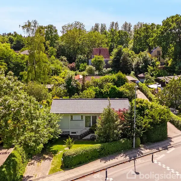 Et grønt hus ligger stille omgivet af tæt løv og frodige træer i et villakvarter. Små veje og haver fylder omgivelserne. Boligsiden-logoet ses i nederste hjørne.