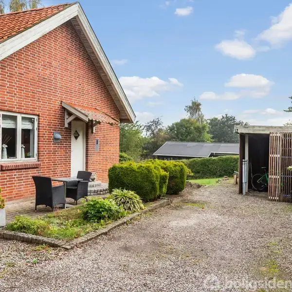 Rødt murstenshus med trekantet tag og hvide vinduesrammer står ved grusbelagt gårdsplads. To sorte lænestole og et lille bord står foran grønne buske ved indgangsdøren, omkranset af træer.