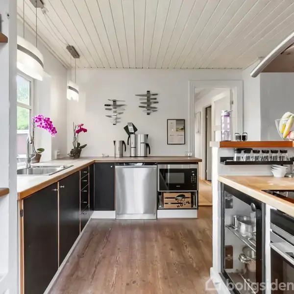 Et moderne køkken har sorte skabe, en træbordplade og apparater som opvaskemaskine og ovn. Der er åbne hylder med glas og flasker og en dekorativ frugtskål på bordet.