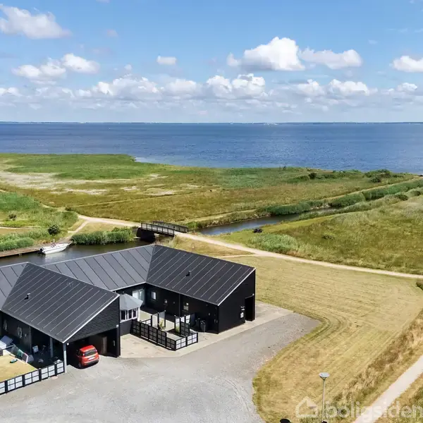 Moderne sorte huse står stille ved en kystlinje, omgivet af grønne græsarealer, under en blå himmel med skyer. I baggrunden ses havet, der strækker sig mod horisonten.