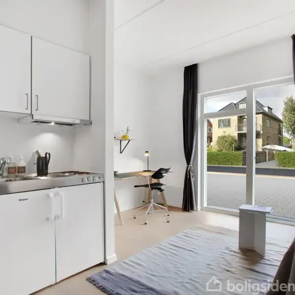 Et lille køkken med hvidt skab og komfur ved siden af en seng, skrivebord med stol, stor vindue. Udenfor ses boligbygninger og grønne buske. En tæppe ligger på gulvet.