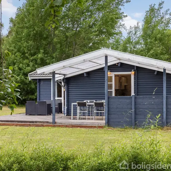 Et blåmalet sommerhus med overdækket terrasse står i en grøn have omkranset af træer. Terrassen har et spisebord med stole og en sofagruppe. Skoven omkranser området.