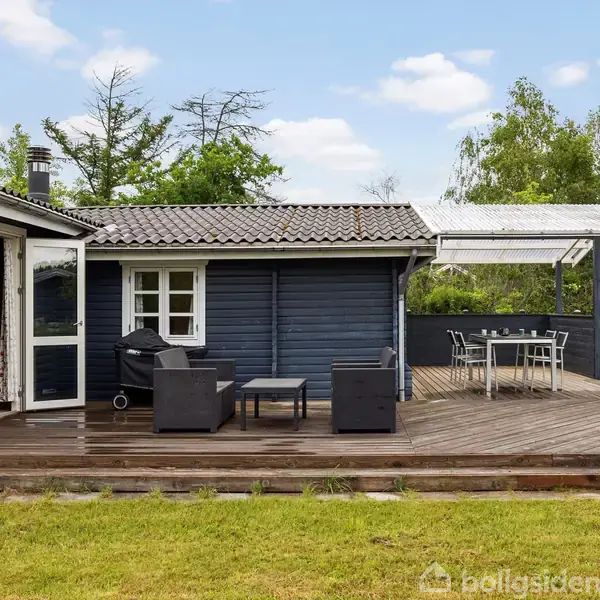 Et sort træhus med terrasse indeholder havemøbler, herunder en grill og siddepladser. Det omgivende område består af en velplejet græsplæne og træer under en klar himmel.