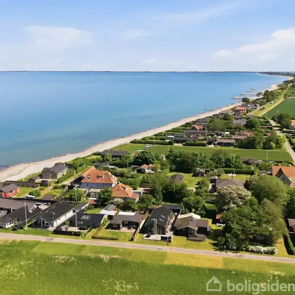 Huse står stille nær en strand langs kysten. Omgivet af grønne områder og en vej, strækker havet sig ud i horisonten under en blå himmel.