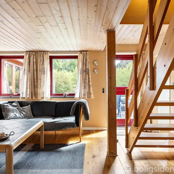 En stue med en trappe i træ fører opad. Sofaer står ved et sofabord på et tæppe. Vinduer med gardiner slipper lys ind, skaber en hyggelig atmosfære.