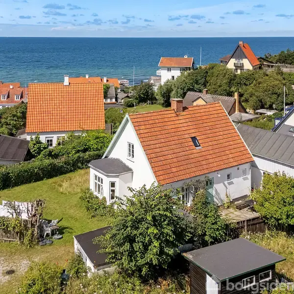 Et hvidt hus med rødt tag står stille omgivet af en have. Det ligger i en kystby med flere huse med havet i baggrunden under en klar blå himmel.