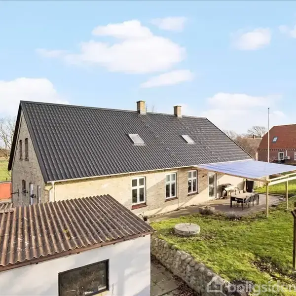 Et hus med sort tag står ved en terrasse med afdækning; en grøn græsplæne omgiver bygningen. I baggrunden ses et rødt hus og åbne marker.