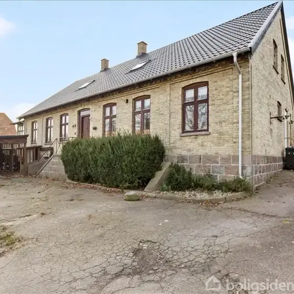 Et ældre, gulstenshus står stille med en gruset gårdsplads foran og grønne buske langs facaden. Baggrund viser et andet hus og en carport med opbevaringsenheder.